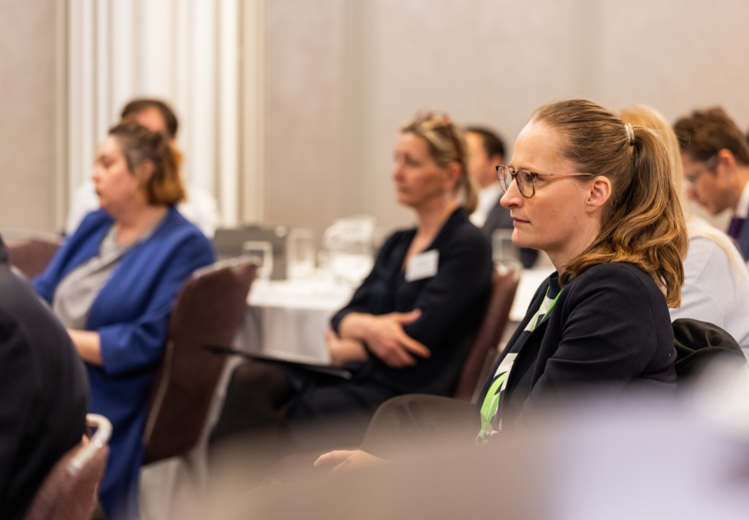 Seated attendees at a conference, listening attentively during a session in a formal meeting room with rows of chairs and tables.